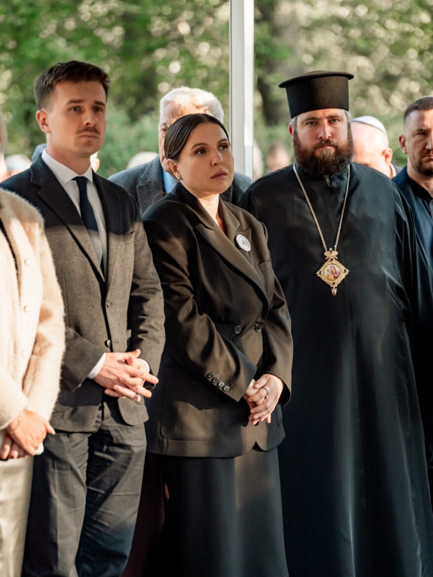 Православний архієрей у камилавці з панагією стоїть поруч із молодою жінкою в чорному та чоловіком у сірому костюмі серед учасників заходу