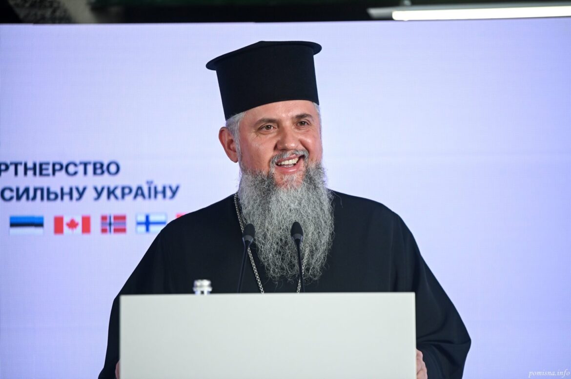 Митрополит Епіфаній у чорному клобуку усміхається, виступаючи за трибуною з двома мікрофонами; позаду екран із написом «Партнерство за сильну Україну» і прапорами